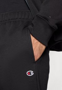 Mão no bolso de umas calças de treino pretas Champion com cós elástico e logo visível da Champion perto do bolso.