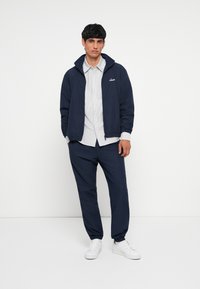 Jūrinės spalvos striukė su užtrauktuku ant apykaklės, derinama su šviesiai pilku dryžuotu marškiniu ir tamsiai mėlynomis siaurėjančiomis kelnėmis. Aprangą užbaigia balti sportiniai bateliai.