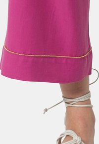 Gonna di stoffa rosa con un accento dorato all'orlo. È visibile il piede di una modella, che indossa sandali beige con lacci attorno alla caviglia.