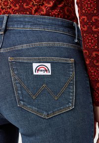Tmavé denimové džínsy s vyšitým dizajnom "W" na zadnej vrecku a nášivkou s logom Wrangler. Čiastočne viditeľný farebný vzorovaný top.