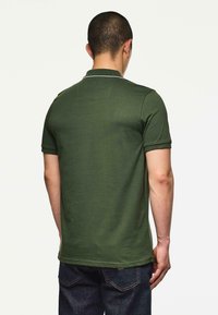 Muž s krátkými vlasy, oblečený v tmavě zeleném polo tričku s krátkým rukávem a tmavě modrých džínách, stojící zády k barevnému pozadí.