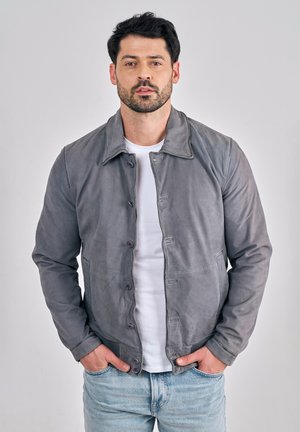 Mann mit dunklem Haar und Bart, der eine graue Lederjacke mit Knöpfen, ein weißes T-Shirt und hellblaue Jeans trägt, Hände in den Taschen, neutraler Hintergrund.