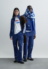 Fatos de treino azuis com detalhes gráficos brancos; mulher com camisa branca, homem com um lenço estampado. Ambos usam sapatos pretos, exibindo um design desportivo.