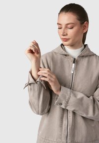 Giacca a vento grigio chiaro con cappuccio realizzata in tessuto morbido, dotata di chiusura frontale con zip, dettagli ai polsini e texture sottile. Il design è minimalista.