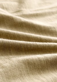 Gros plan d’un tissu tissé beige doux avec une texture subtilement côtelée et des plis doux créant des variations de lumière et d’ombre.