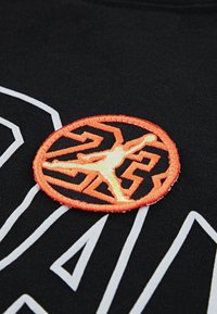 Μαύρο ύφασμα με λευκό γραφικό σχέδιο. Διαθέτει στρογγυλό, πορτοκαλί κεντητό λογότυπο με σιλουέτα και γεωμετρικά μοτίβα.