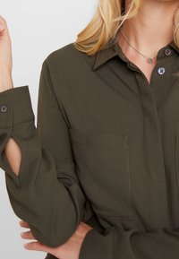 Olivově zelená košile s dlouhým rukávem, širokým límcem a knoflíky, dvěma náprsními kapsami a stehenními akcenty na rukávech. Hladká textura látky.
