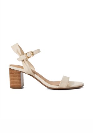 Beige heeled sandal with a wooden block heel, satin straps, and an adjustable buckle. Features a knotted accent on the ankle strap.