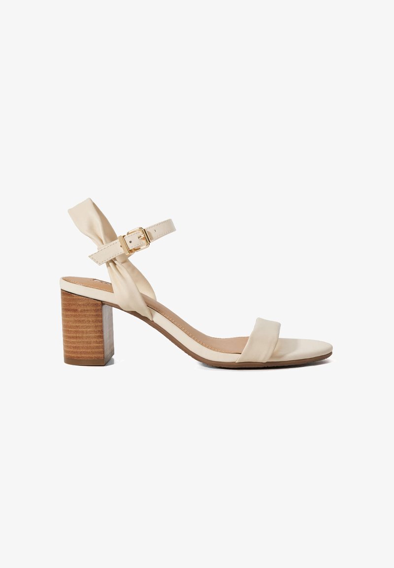 Beige heeled sandal with a wooden block heel, satin straps, and an adjustable buckle. Features a knotted accent on the ankle strap.