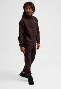 Mężczyzna idący w ciemnobrązowej bluzie z zamkiem i dopasowanych spodniach, w czarnych trampkach, na jednolitym białym tle.