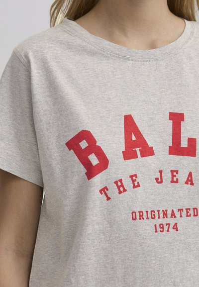 Pelēka kokvilnas T-kreklā ir lieli sarkani burti ar uzrakstu "BAL THE JEAN ORIGINATED 1974". T-krekla dizainā ir apaļa kakla izgriezne un īsas piedurknes.