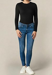 Personne portant un haut à manches longues noir, un jean skinny bleu et des baskets blanches, debout devant un fond beige uni.