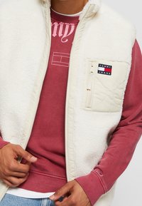 Kremowa kamizelka z polaru z zamkiem z przodu, z pikowaną kieszenią i naszywką z logo. Nosi się ją na bordowej bluzie z prążkowanymi akcentami.