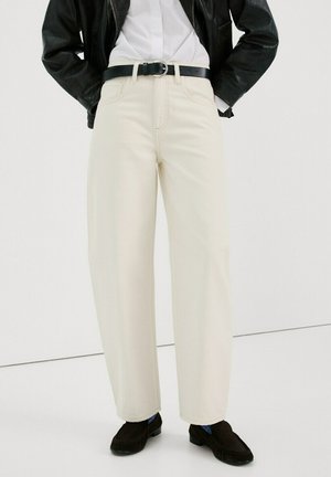 Persona con pantalones crema de talle alto y pierna ancha, cinturón negro, camisa blanca metida, chaqueta negra y mocasines marrón oscuro.