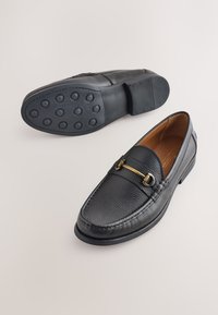 Mocassins en cuir noir avec une finition texturée, des détails en métal doré, et une semelle en caoutchouc présentant un motif de crampons circulaires.