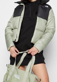 Jasnozielona kurtka puchowa z czarnymi panelami na ramionach, z zamkiem z przodu. Trzyma zieloną torbę typu tote z regulowanymi paskami i detalami z logo.