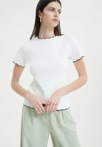 Biely top s krátkym rukávom a čiernym volánovým lemovaním okolo výstrihu a lemov, skombinovaný so svetlozelenými nohavicami. Hladká textúra a priliehavý strih.