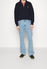 Felpa blu navy con zip e polsini a coste, jeans svasati azzurri chiari e scarpe nere con suola color cammello. La felpa presenta un logo ricamato.