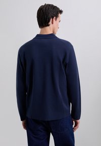 Pulover polo cu mâneci lungi de culoare albastru marin, cu un guler simplu, textură moale și croială lejeră, văzut din spate. Purtați cu blugi negri.