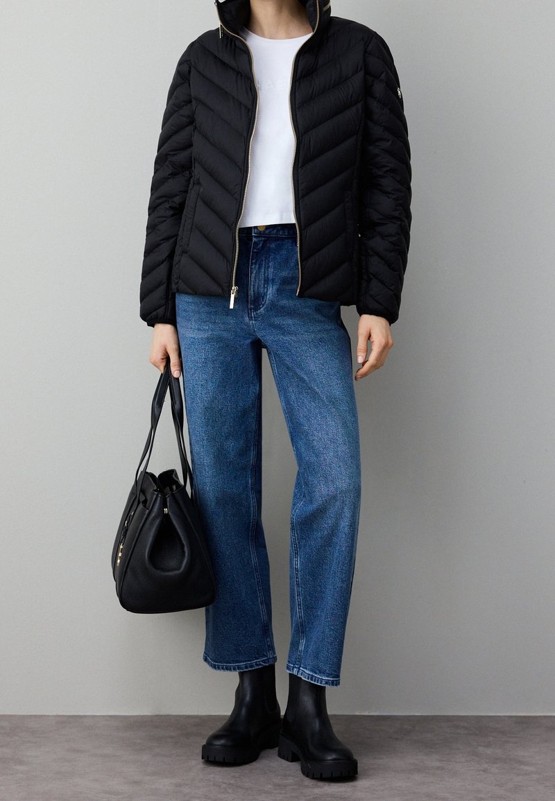 Doudoune noire à motif chevron, t-shirt blanc, jean taille haute bleu et bottes noires épaisses, tenant un sac à main noir.