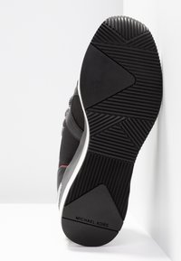 Podeszwa czarnych sneakersów Michael Kors z teksturowanym wzorem przeciwpoślizgowym i białą podeszwą środkową na białym tle.