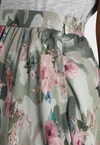 Jupe à motif floral avec un fond camouflage, présentant une taille froncée et une fermeture éclair discrète. Les couleurs incluent le rose, le vert et le gris.