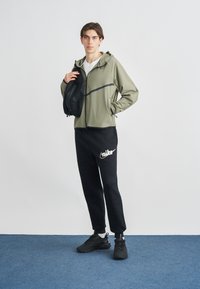 Jasnooliwkowa kurtka na zamek błyskawiczny z czarnym paskiem, czarne spodnie dresowe z białym logo, czarne buty i czarna torba. Gładkie tło.