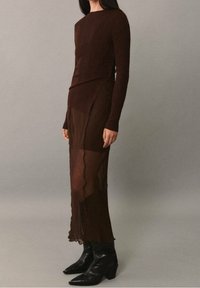 Haut marron à manches longues côtelé avec un petit décolleté, superposé sur une jupe marron transparente à ourlet ondulé, associé à des bottines noires pointues.