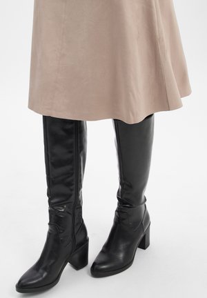 Zwarte leren kniehoge laarzen met blokhakken gedragen onder een beige suède rok tegen een witte achtergrond.