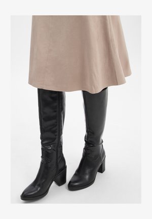 Schwarze knielange Lederstiefel mit Blockabsätzen, getragen unter einem beigen Wildlederrock vor einem weißen Hintergrund.