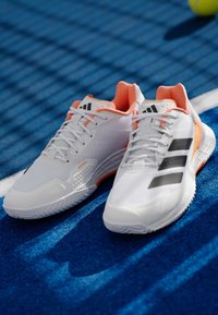 Λευκά αθλητικά παπούτσια με ύφασμα από δίχτυ, πορτοκαλί λεπτομέρειες, τρεις μαύρες ρίγες και υφή στην σόλα, σε μια ζωηρή μπλε επιφάνεια.