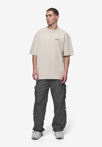 Beige ylittävä t-paita lyhyillä hihoilla ja pieni musta logo. Tummanharmaat cargo-housut, joissa on useita taskuja ja säädettävät hihnat. Rentouttavat lenkkarit.