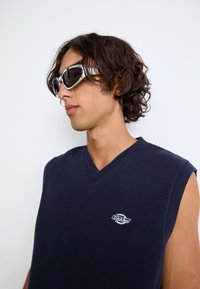 Jersey sin mangas azul marino con escote en V y un pequeño logo blanco en el pecho. El modelo lleva gafas de sol plateadas con lentes negros.