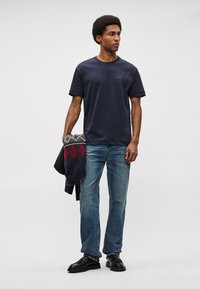 Ναυτικό μπλε βαμβακερό t-shirt με κοντά μανίκια, συνδυασμένο με ανοιχτό γαλάζιο τζιν παντελόνι. Κρατάει μια σκούρα κουβέρτα με κόκκινες και γεωμετρικές σχέσεις.