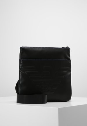 Schwarze Leder-Umhängetasche mit eingeprägtem Logo auf der Vorderseite, Reißverschluss und verstellbarem Riemen, positioniert auf weißer Oberfläche vor grauem Hintergrund.