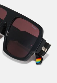 Ochelari de soare de dimensiuni mari, de culoare neagră, cu lentile roșii închise. Designul cu ramă pătrată are un accent colorat lângă balama și branding Polaroid.