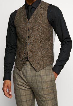 Mężczyzna ubrany w brązową kamizelkę w jodełkę zapinaną na guziki, na czarnej koszuli i krawacie, w połączeniu z beżowymi spodniami w kratę, z ręką w kieszeni.