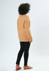 Lysebrunt oversized sweater med lange ærmer, parret med slanke sorte leggings og lysebrune blokhælede mules, vist bagfra.