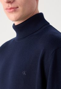 Navyblauer Rollkragenpullover aus gestricktem Material. Mit einem kleinen, geprägten Logo auf der Brust, einem taillierten Kragen und glatter Struktur.