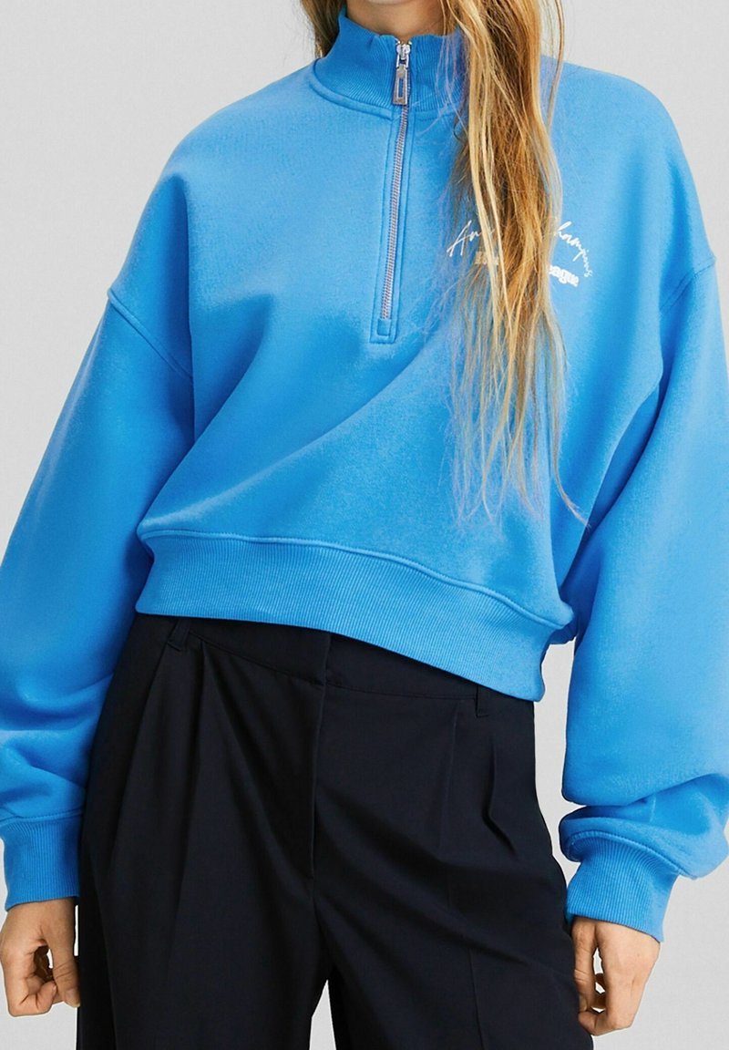 Sudadera azul corta con cuello alto y cremallera, puños y cintura acanalados. Presenta un bordado sutil del logo en el frente. Textura suave.