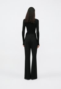 Mulher com cabelo encaracolado, vestindo um top preto de mangas longas e calças largas pretas de cintura alta, voltada para longe em frente a um fundo simples.