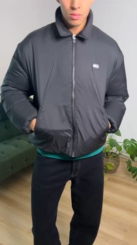 Νεαρός άνδρας με μαύρο μπουφάν με φερμουάρ και τα χέρια στις τσέπες, στέκεται σε εσωτερικό χώρο κοντά σε πράσινο καναπέ και γλαστράκι.