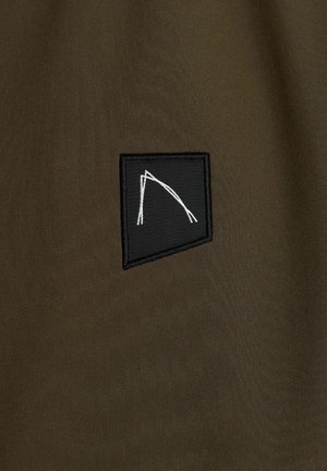 Oliivivihreä kangas, jossa on mattapinta, ja musta neliömäinen applikaatio, johon on brodeerattu valkoinen abstrakti muoto, joka koostuu risteävistä viivoista.