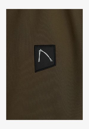 Oliivivihreä kangas, jossa on mattapinta, ja musta neliömäinen applikaatio, johon on brodeerattu valkoinen abstrakti muoto, joka koostuu risteävistä viivoista.