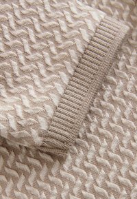 Suéter de punto con un patrón texturizado en beige y líneas onduladas; puños acanalados en un tono más claro que complementa. Tejido suave y ligero.