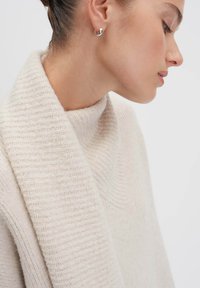 Donna che indossa un morbido maglione lavorato a maglia color crema con un ampio colletto, mostrata di profilo con un orecchino a cerchio in argento all'orecchio.