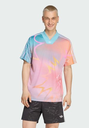 Nasmejana mlada osoba s kratkom plavom kosom, odjevena u Adidas dres s pastelnim gradijentom i crne šarene Adidas kratke hlače na jednobojnoj pozadini.