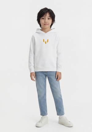 Kind met kort zwart haar, draagt een witte hoodie met oranje logo, lichtblauwe spijkerbroek, witte sokken en witte sneakers, staande tegen een witte achtergrond.