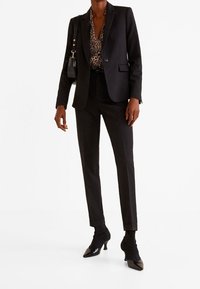 Personne portant un costume noir sur mesure avec un chemisier à imprimé léopard, des bottines à talons pointues noires et un sac  à bandoulière noir.