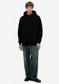 Sudadera negra de gran tamaño con capucha ajustable, combinada con jeans de mezclilla de corte holgado en un tono azul desgastado y zapatos de cuero negros.